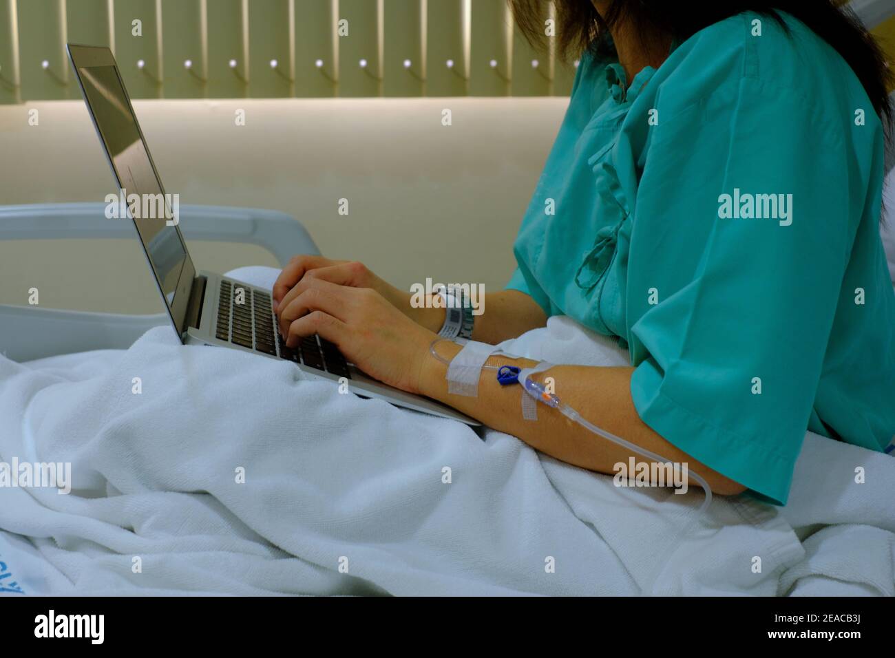 Un colpo di mezzo corpo di una donna d'affari lavoraholic seduta sul suo letto dell'ospedale con iniezione di IV nel suo braccio, recuperando dall'ambulatorio mentre usando il suo lapt Foto Stock