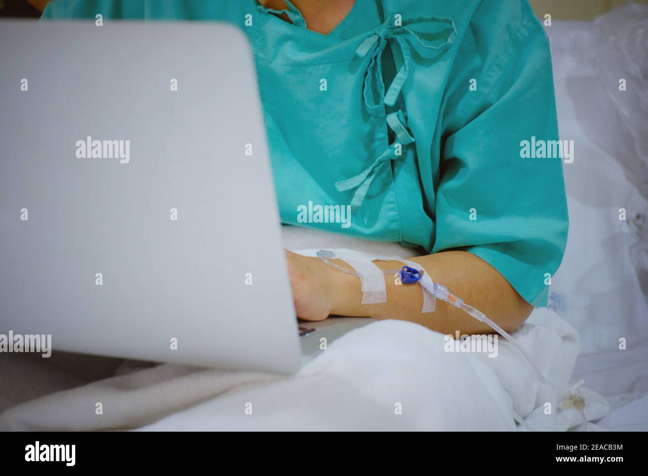 Un colpo di mezzo corpo di una donna d'affari lavoraholic seduta sul suo letto dell'ospedale con iniezione di IV nel suo braccio, recuperando dall'ambulatorio mentre usando il suo lapt Foto Stock