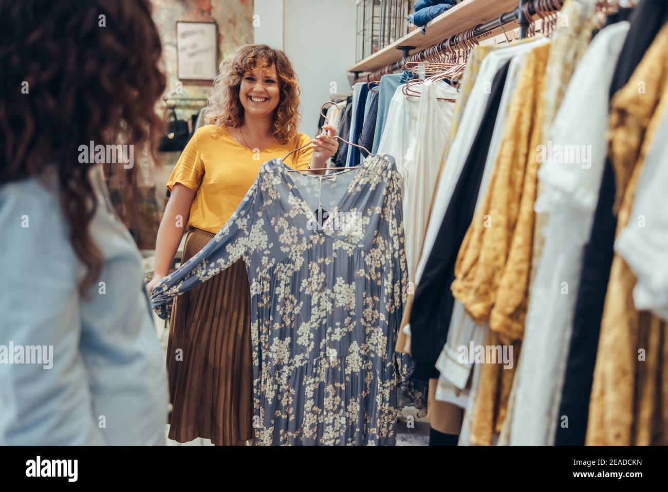 Donna che tiene un abito e lo mostra alla cliente femminile nel suo negozio. Il proprietario del negozio di abbigliamento aiuta un cliente in negozio. Foto Stock