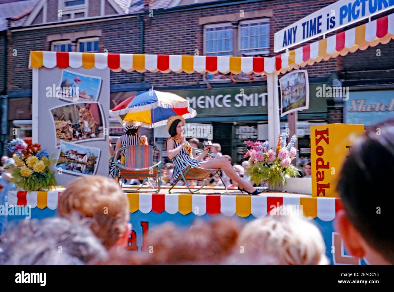 Un float parata per film, processing e stampe Kodak alla sfilata annuale del Carnevale di Whitsun, Watford, Hertfordshire, Inghilterra, UK 1965. Una donna in un mini abito seduta su una sedia da spiaggia sta tenendo una macchina fotografica Kodak. Una scatola gigante di pellicola Kodak è inoltre sul galleggiante della parata. Questa immagine proviene da un vecchio lucido a colori amatoriale Kodak da 35 mm. Foto Stock