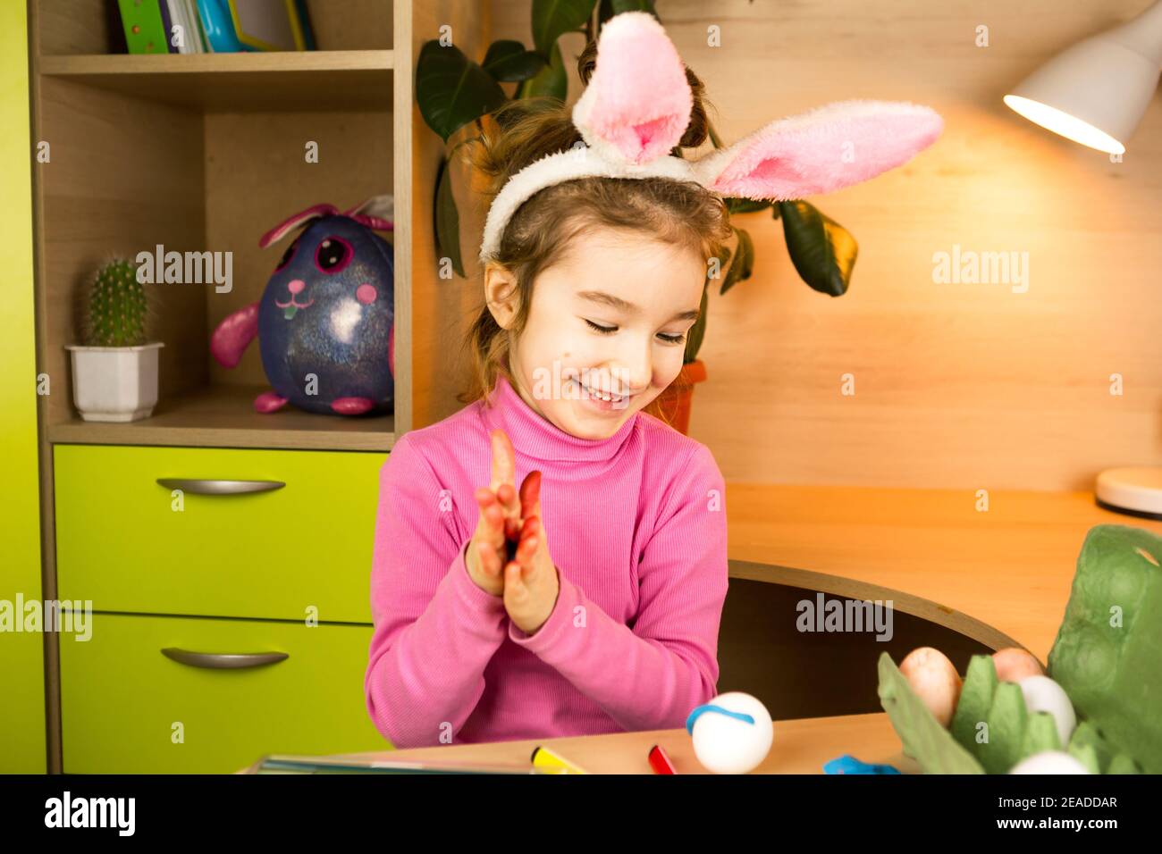 Una ragazza nelle orecchie di un coniglietto pasquale dipinge le uova con una penna di feltro-punta nell'interno domestico. Artigianato, preparazione per una festa religiosa, un vassoio con e. Foto Stock