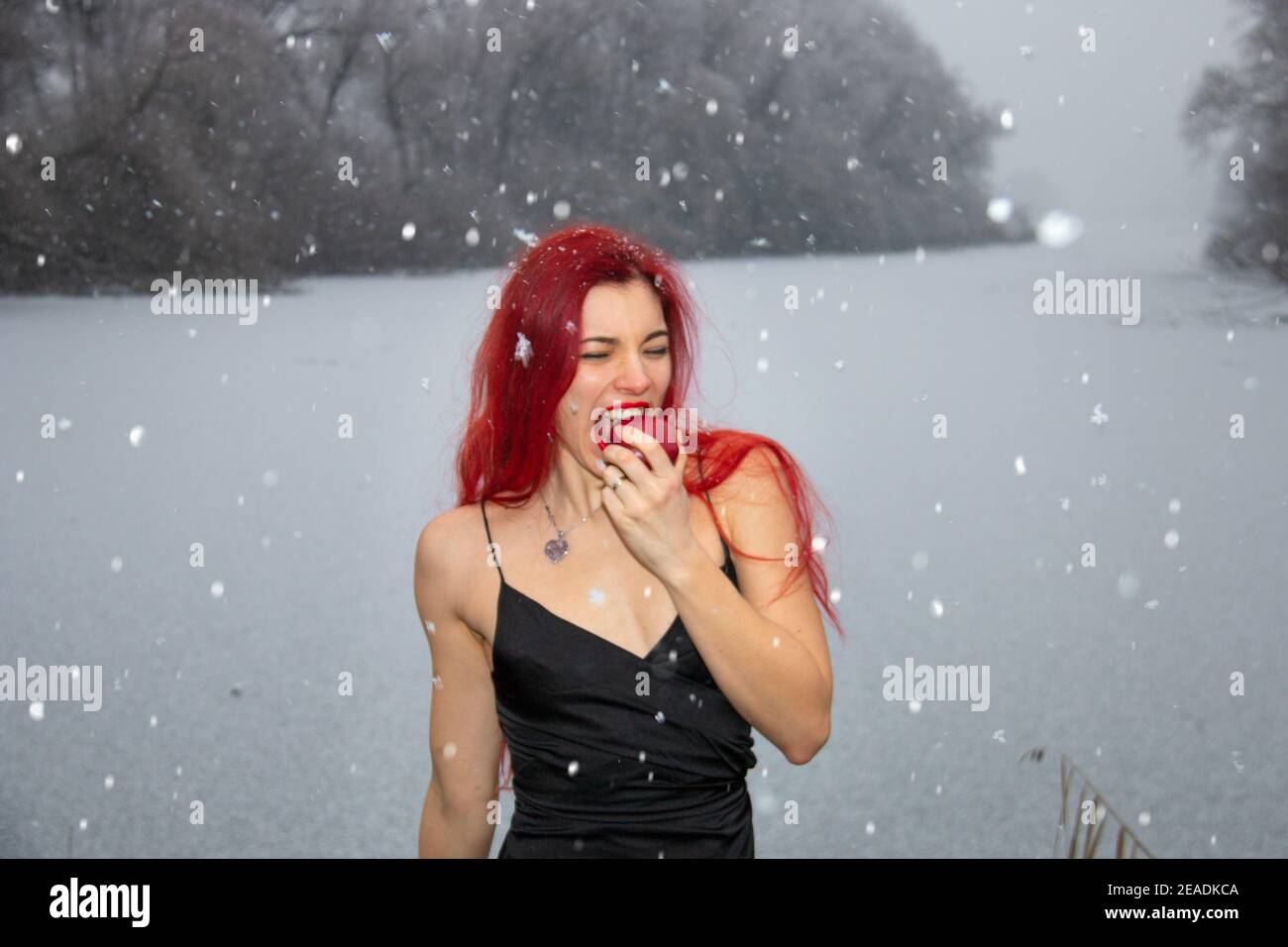 La donna dei capelli rossi sta mangiando una mela mentre la doccia della neve all'aperto nella natura in inverno freddo lago ghiacciato Foto Stock