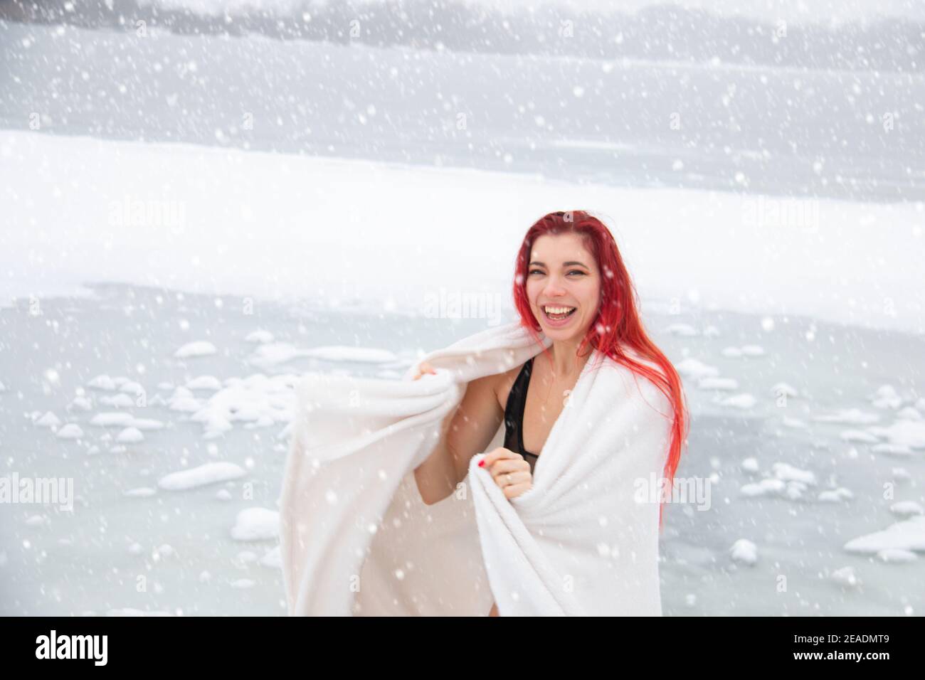 Ritratto invernale innevato di felice donna sorridente in caduta di neve all'esterno in un costume da bagno, avvolto in un asciugamano presso il lago di nuoto in acque aperte Foto Stock