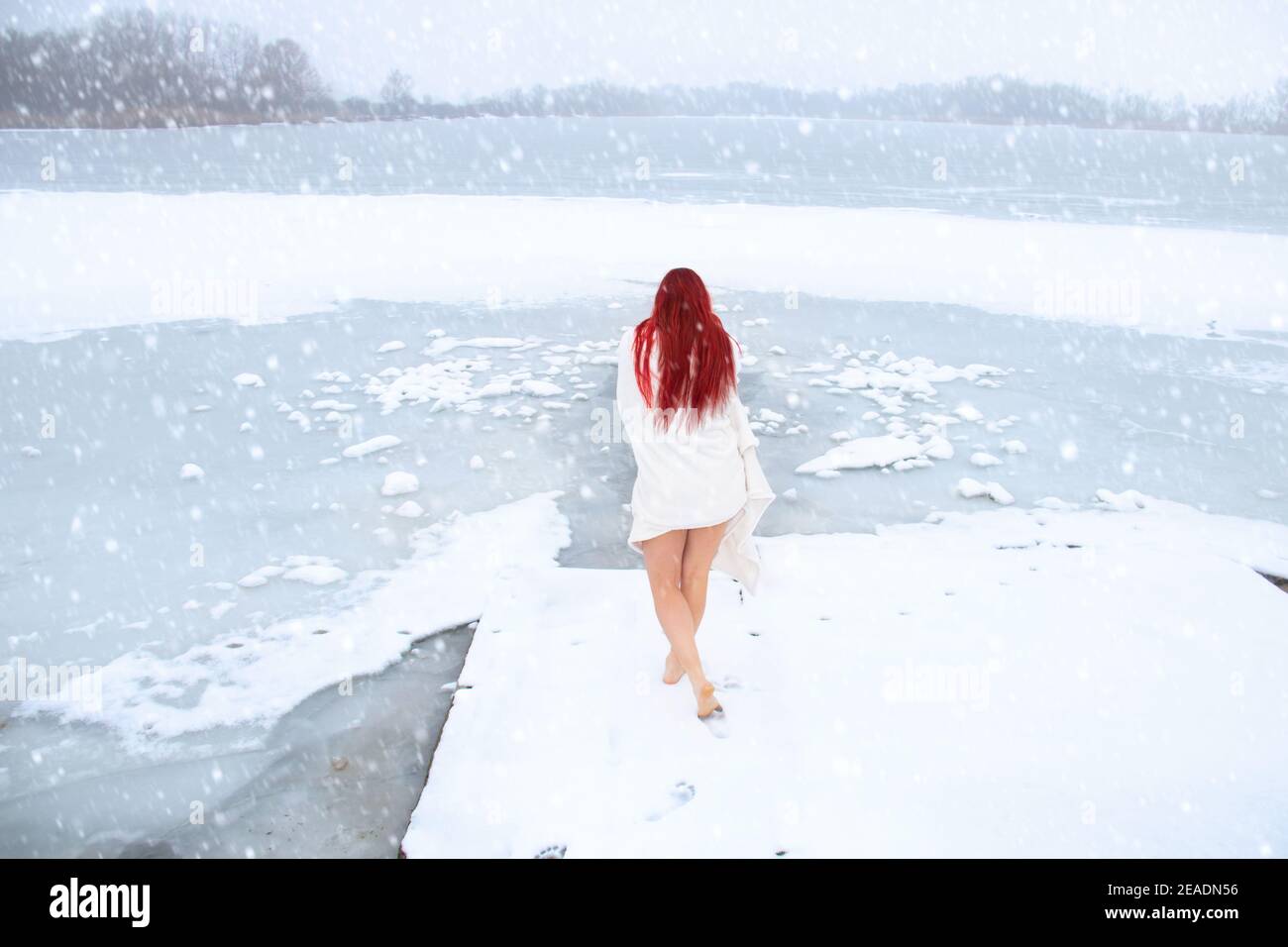 Donna con vista posteriore con lunghi capelli rossi e belle gambe camminando all'esterno a piedi nudi sulla neve fino al lago ghiacciato per il bagno di ghiaccio naturale mentre nevica Foto Stock