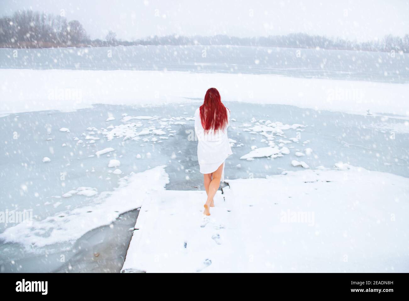 Bella donna testa rossa con capelli lunghi vista posteriore avvolta solo in un tovagliolo bianco polare che cammina con le gambe nude la neve verso un lago gelido aperto Foto Stock