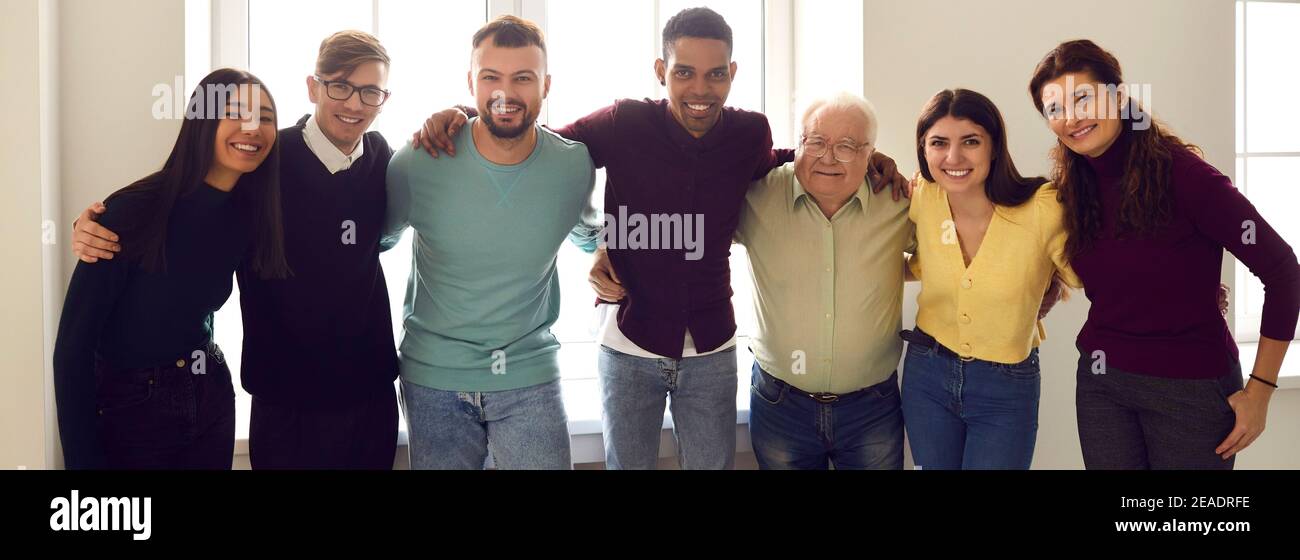 Ritratto di gruppo di un gruppo di persone diverse che si abbracciano l'un l'altro in piedi accanto alla finestra insieme. Foto Stock