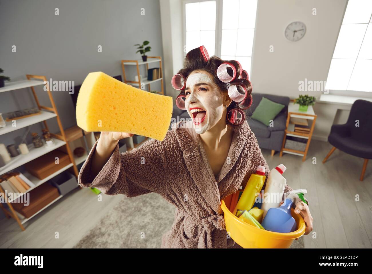 Divertente giovane casalinga pazza in arricciacapelli e cura della pelle faccia maschera che riordinare la sua casa Foto Stock
