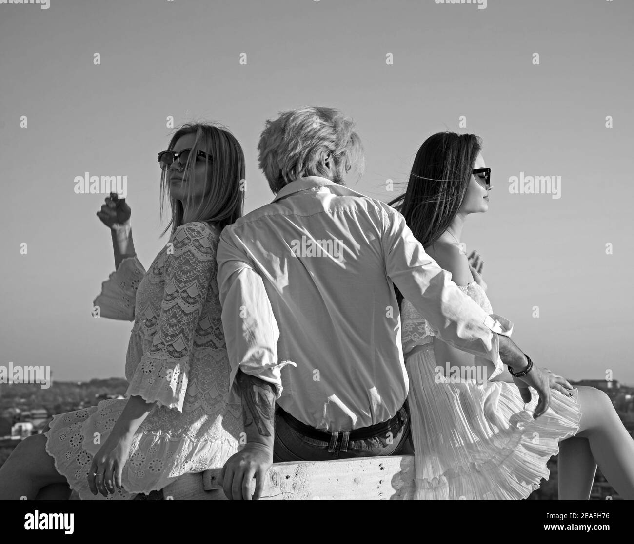 Due graziose ragazze e un uomo brutale che si siedono tra loro guardando in lontananza sullo sfondo del cielo azzurro. Foto Stock