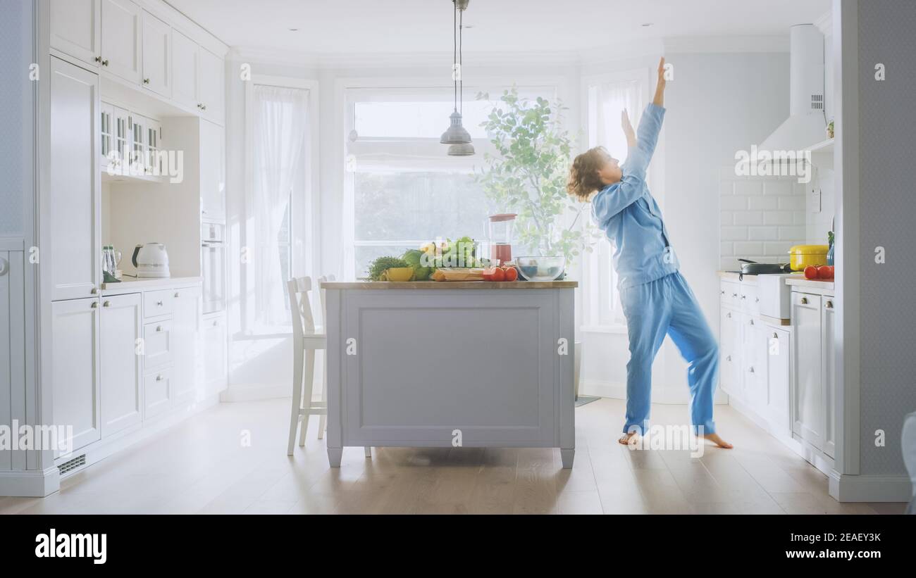 Energico giovane uomo divertente con lunghi capelli che ballano in cucina mentre indossa le Pajama blu. Area cucina moderna di colore bianco luminoso con verde sano Foto Stock