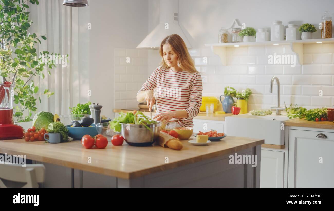 Giovane femmina in ponticello a strisce sta facendo una sana insalata biologica in una moderna cucina soleggiato. La donna autentica sta tagliando un cavolfiore con un coltello Foto Stock