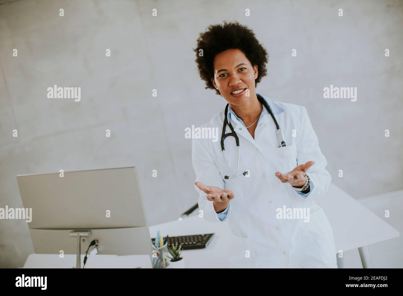 Donna Africana americana medico indossando camice bianco con stetoscopio in piedi dalla scrivania in ufficio Foto Stock