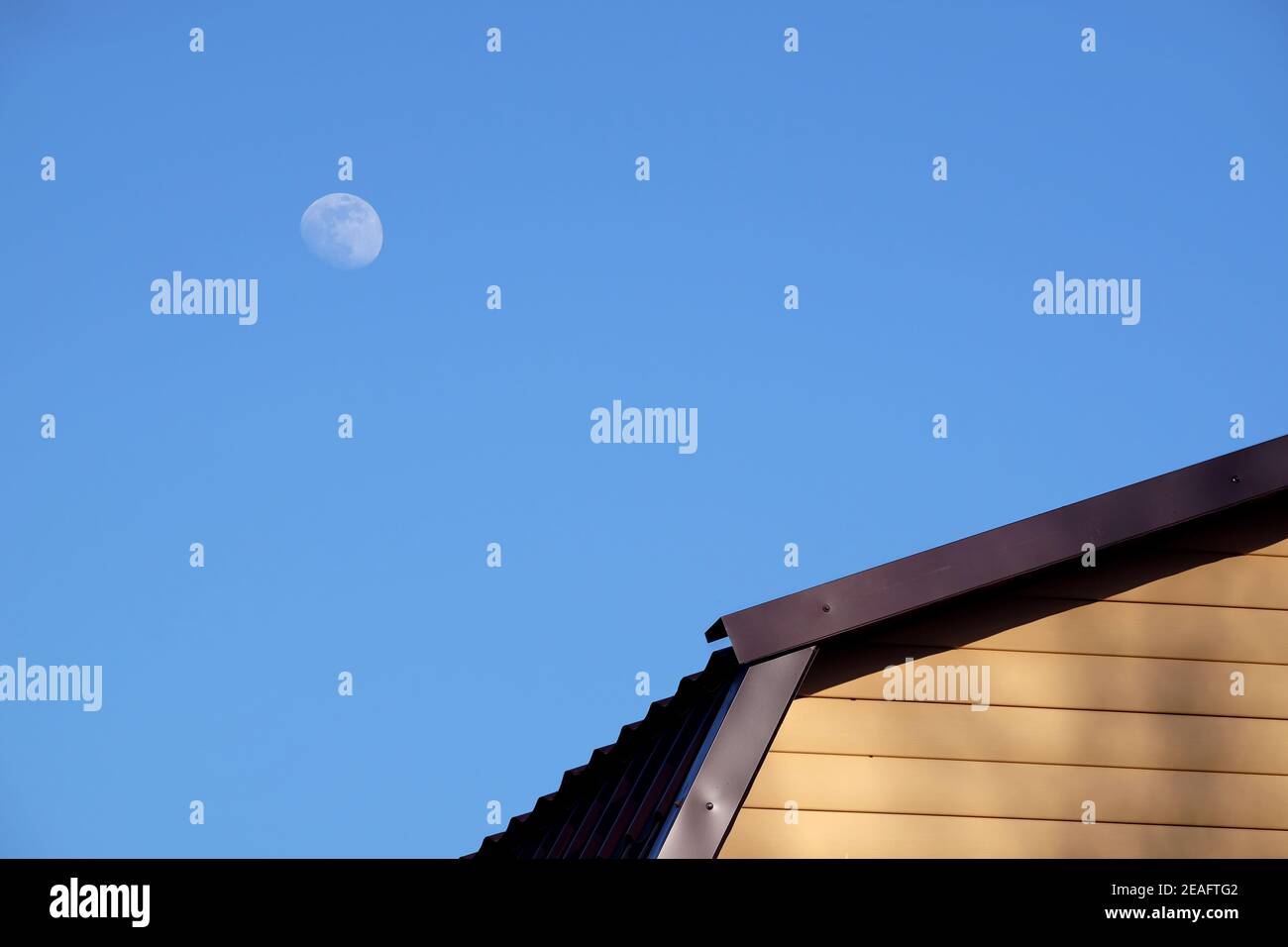 Parte della parete rurale della casa coperta con il lato giallo e. tetto in metallo marrone e luna rotonda su blu chiaro senza nuvole cielo sulla soleggiato giorno vista frontale closeup Foto Stock