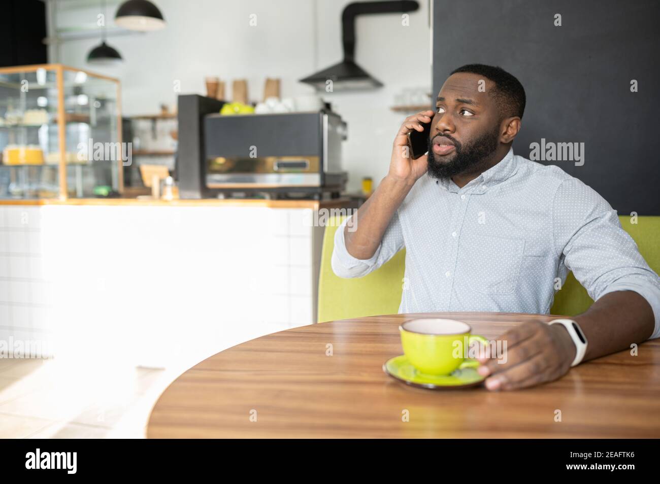 Un ragazzo afro-americano perplesso è seduto nel caffè con una tazza di caffè e ha una conversazione telefonica seria, un uomo multirazziale che parla sullo smartphone Foto Stock