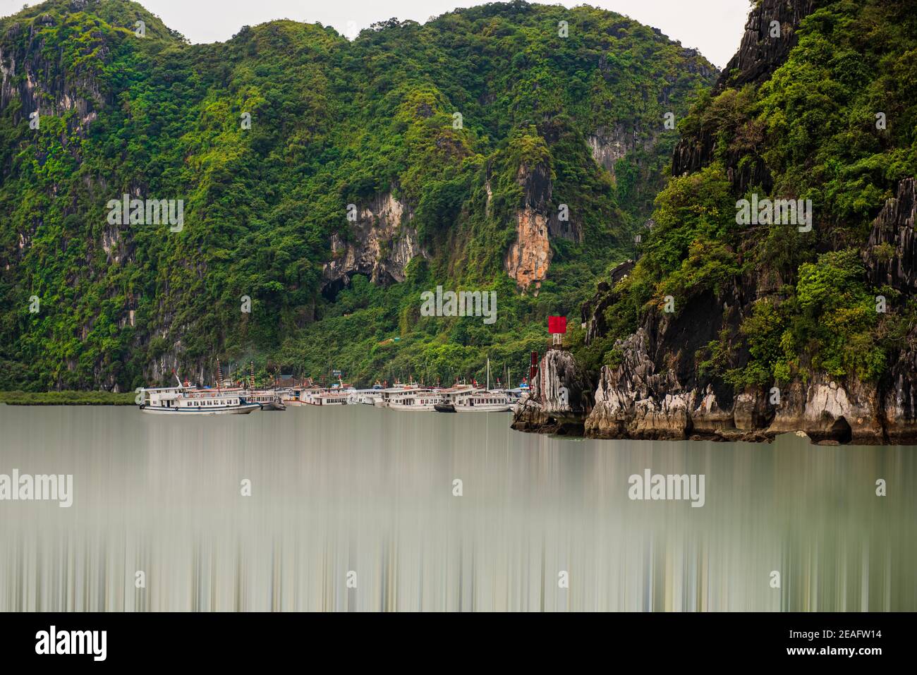 Foto delle barche ormeggiate alla base di un'isola di montagna stonata nelle acque levigate della Baia di ha Long. Foto Stock