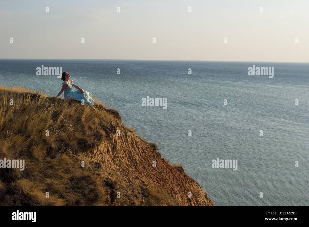 Una ragazza in un vestito lungo si siede su una scogliera vicino al mare Foto Stock