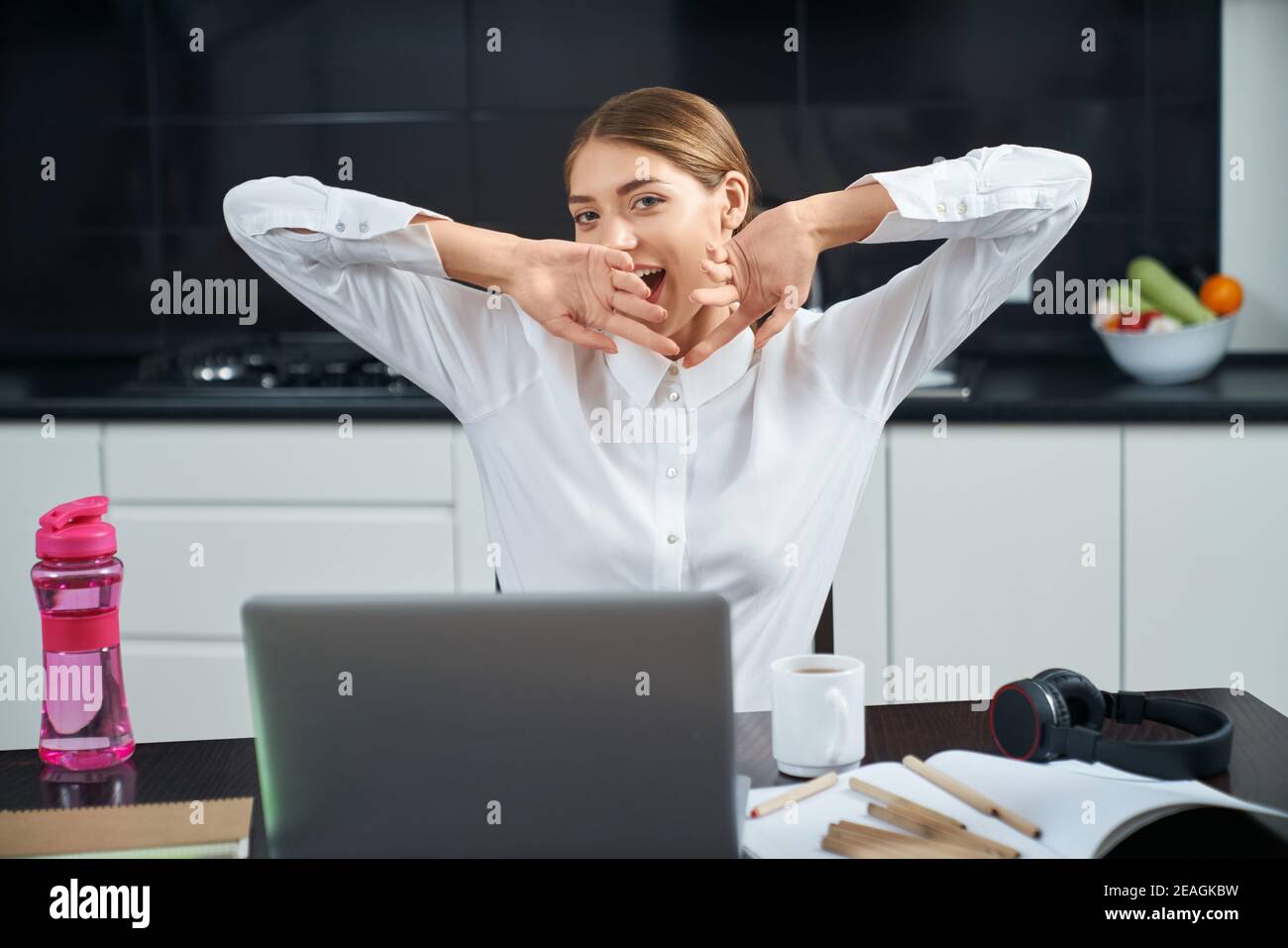 Bella giovane donna seduta al tavolo con il portatile aperto, stretching e sbadigliare. Piacevole sensazione di stanchezza femminile dopo una lunga giornata di lavoro a casa. Foto Stock