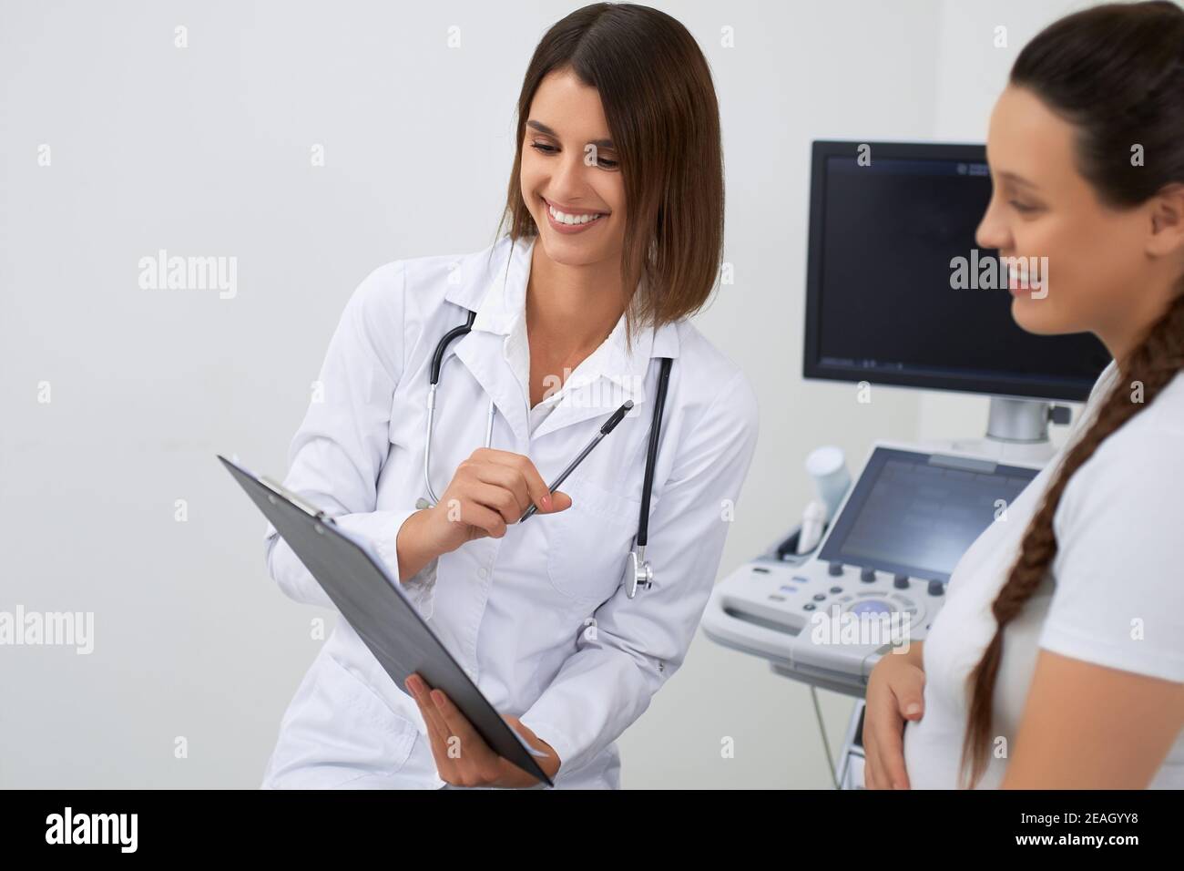 Abile ginecologa femminile in camice di laboratorio che parla con la donna incinta durante l'appuntamento. Dottore che scrive in clipboard tutte le informazioni sul futuro Foto Stock