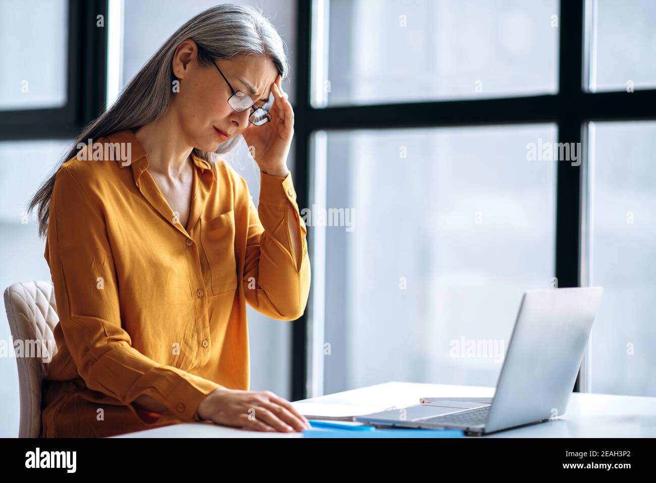 Triste asiatico medio invecchiato grigio-capelli donna d'affari o tutor femminile, è stanco di lavoro straordinario on-line, o di condurre insegnamento a distanza, chiuso gli occhi, mal di testa, bisogno di un riposo Foto Stock