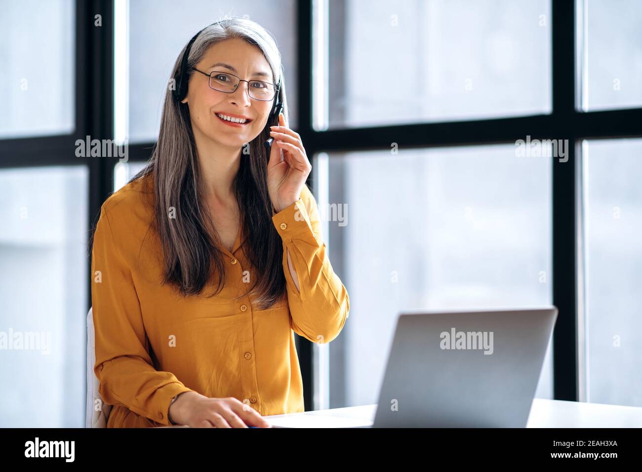 Amichevole asiatico maturo grigio-capelli donna di affari o tutor, in abbigliamento casual elegante, cuffie e occhiali sta lavorando online, o condurre insegnamento a distanza, utilizzando il laptop sul luogo di lavoro, guarda a. Foto Stock