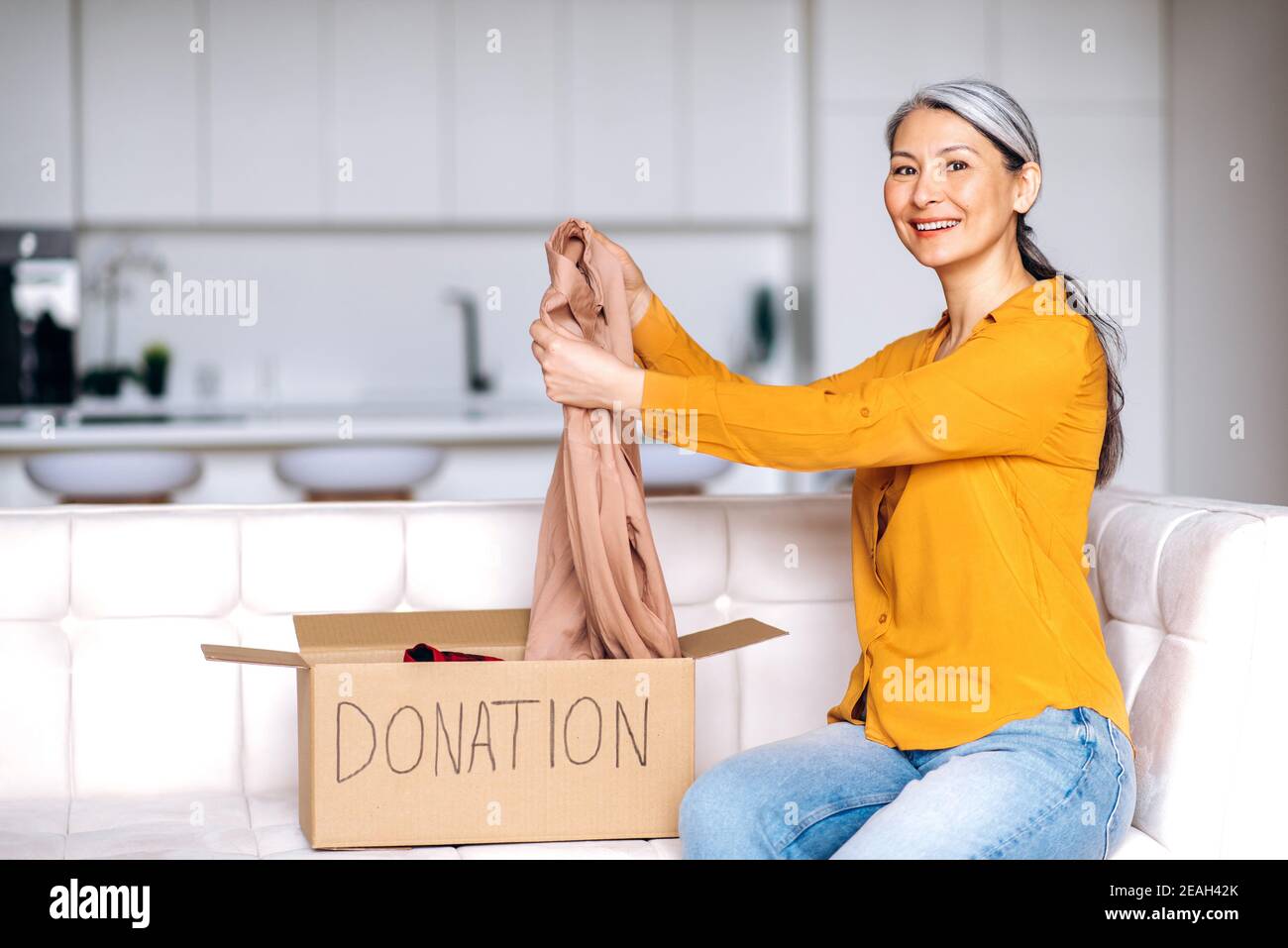 Felice donna anziana con capelli grigi gioisce della donazione, seduta a casa sul divano smalti la scatola di donazione con di vestiti, per il riutilizzo. Aiutare le persone povere e bisognose Foto Stock