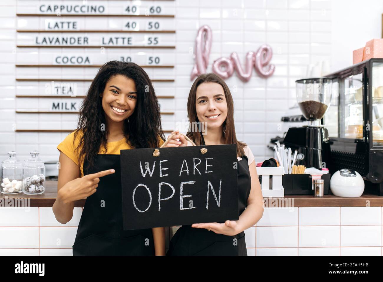 Due cameriere afroamericane e caucasiche soddisfatte una caffetteria o un ristorante, si alza tenendo un cartello SIAMO APERTI vicino al banco bar e sorridente amichevole Foto Stock