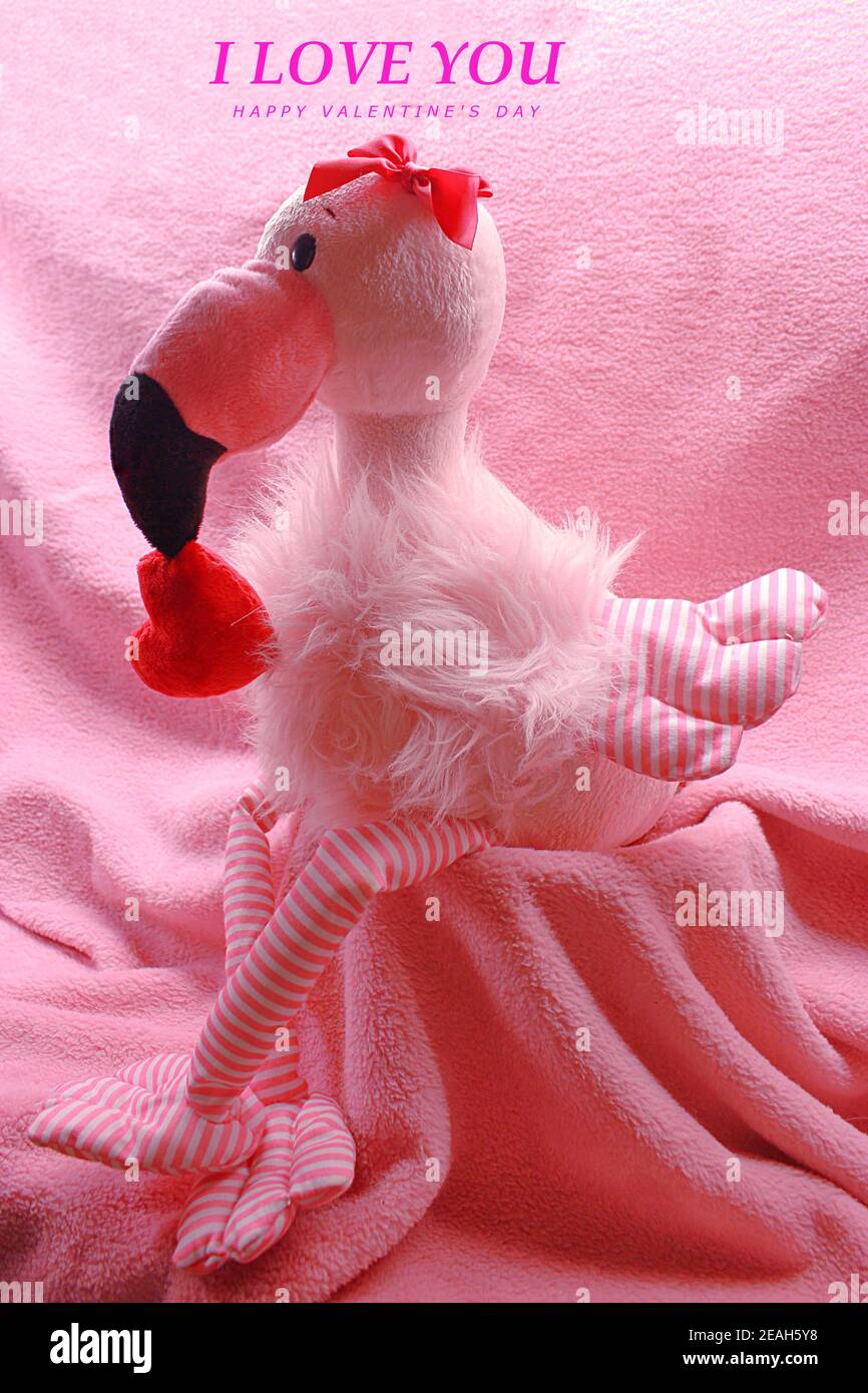 Ti amo e felice giorno di San Valentino scritto sopra a. flamingo seduto su una coperta rosa che tiene un cuore dentro la bocca e il nastro rosso sulla testa Foto Stock
