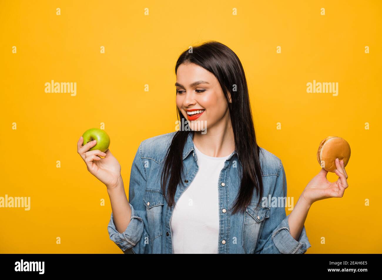 Fast food o cibi sani. Caucasica attraente bruna ragazza tiene una mela in una mano e nell'altra hamburger, facendo una scelta, in piedi su isolato sfondo arancione Foto Stock