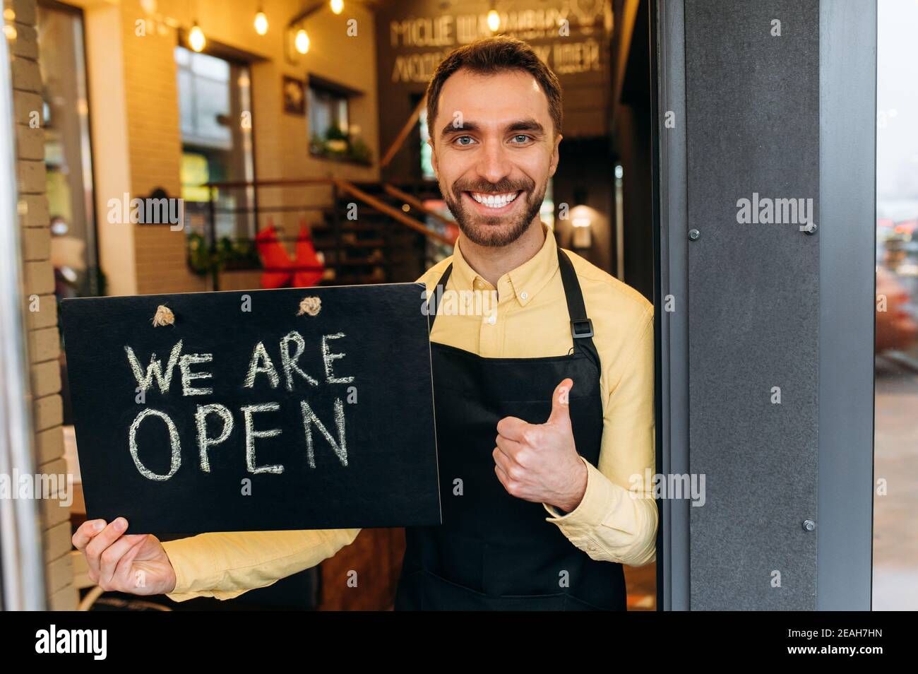 Benvenuto, siamo aperti. Ritratto di gioioso cameriere maschile caucasico, barista o proprietario in piedi alla porta di un ristorante o caffè, tiene un cartello APERTO, e sorriso amichevole. Supportare il concetto di piccola impresa Foto Stock