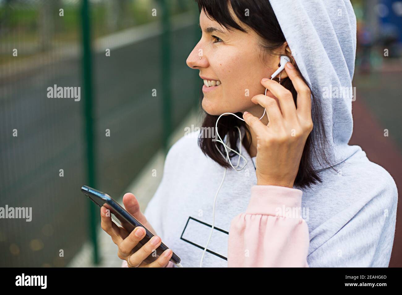 Una donna caucasica utilizza uno smartphone con cuffie cablate e un auricolare. Telefono in mano, filo bianco nell'orecchio, etichetta sulla felpa con cappuccio. Ascolta la musica tramite Foto Stock