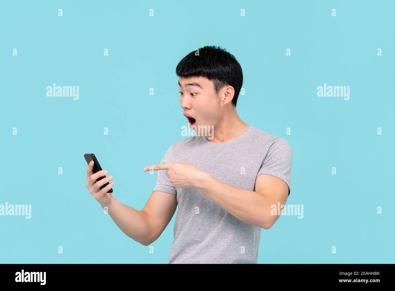 Scioccato eccitato giovane asiatico che anelava e punta al mobile telefono isolato su sfondo azzurro Foto Stock