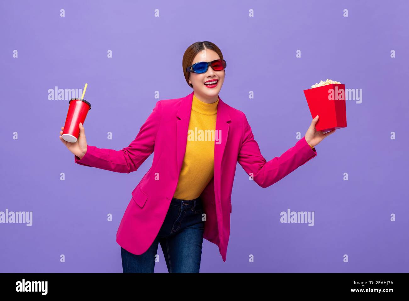 Giovane donna asiatica che indossa occhiali 3D godendo di guardare film con bere e popcorn in mani su sfondo viola Foto Stock