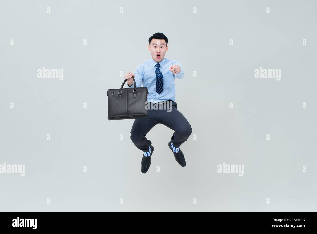 Scioccato eccitato giovane uomo d'affari asiatico con valigia che salta a metà -aria e dito puntato verso la telecamera isolato su grigio chiaro sfondo Foto Stock