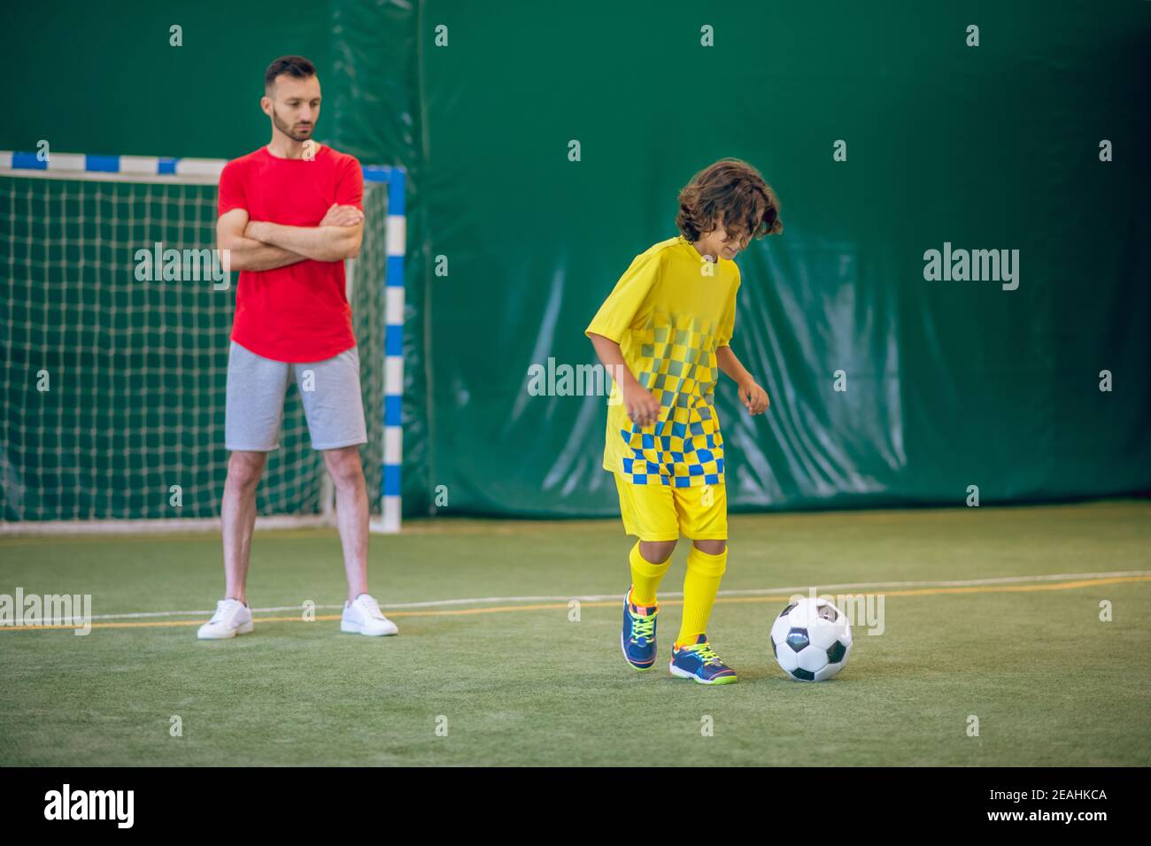 Ragazzo carino in uniforme gialla che gioca a calcio, il suo allenatore che lo guarda Foto Stock