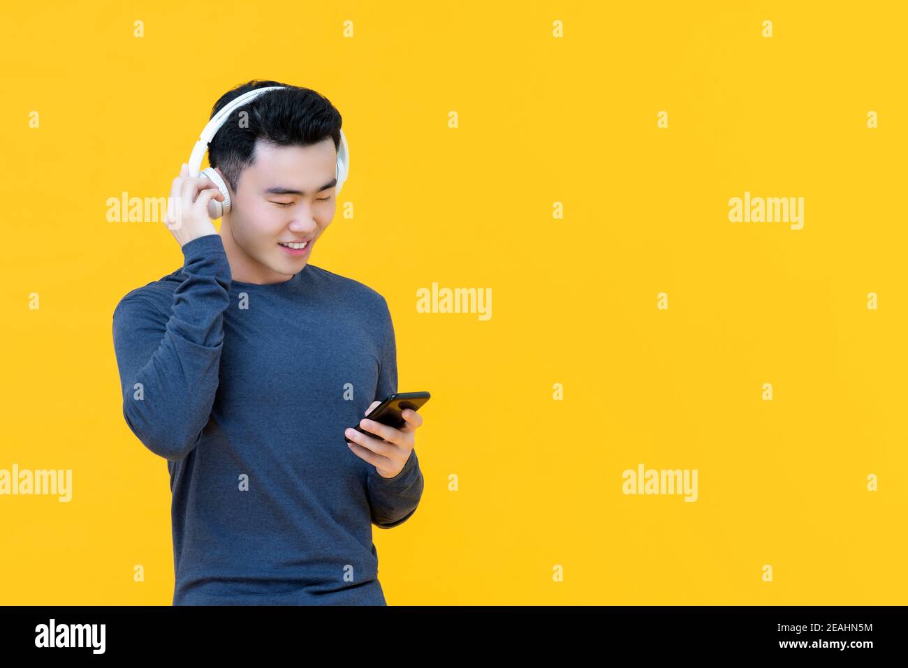 Giovane uomo asiatico felice che indossa le cuffie per ascoltare musica in streaming da telefono sarrt isolato su sfondo giallo Foto Stock