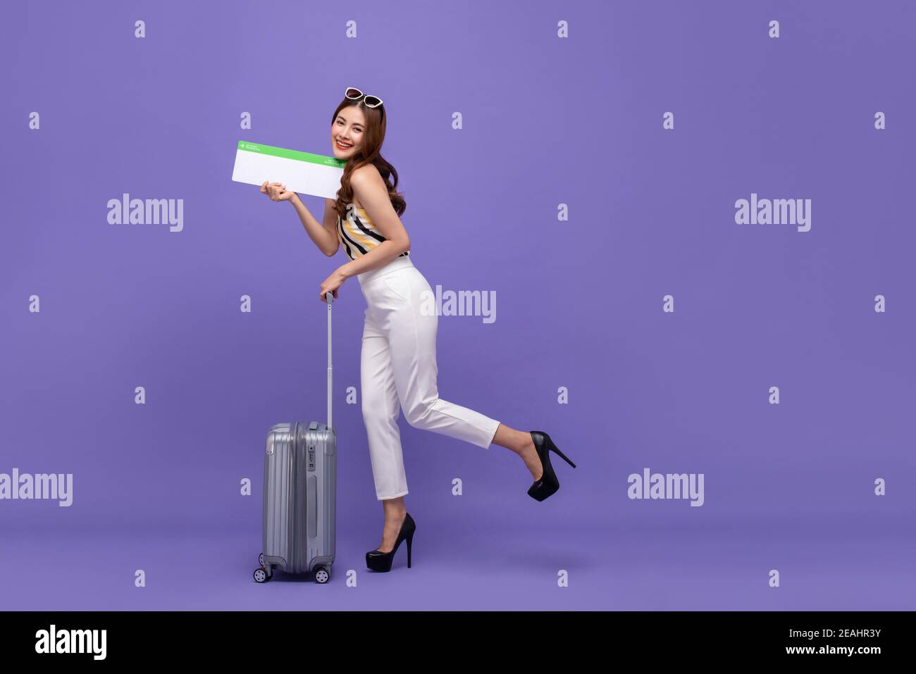 Giovane bella donna asiatica turista in cerca felice ed eccitata sorridente e camminando con i bagagli e tenendo in mano il biglietto aereo isolato sfondo viola per Foto Stock