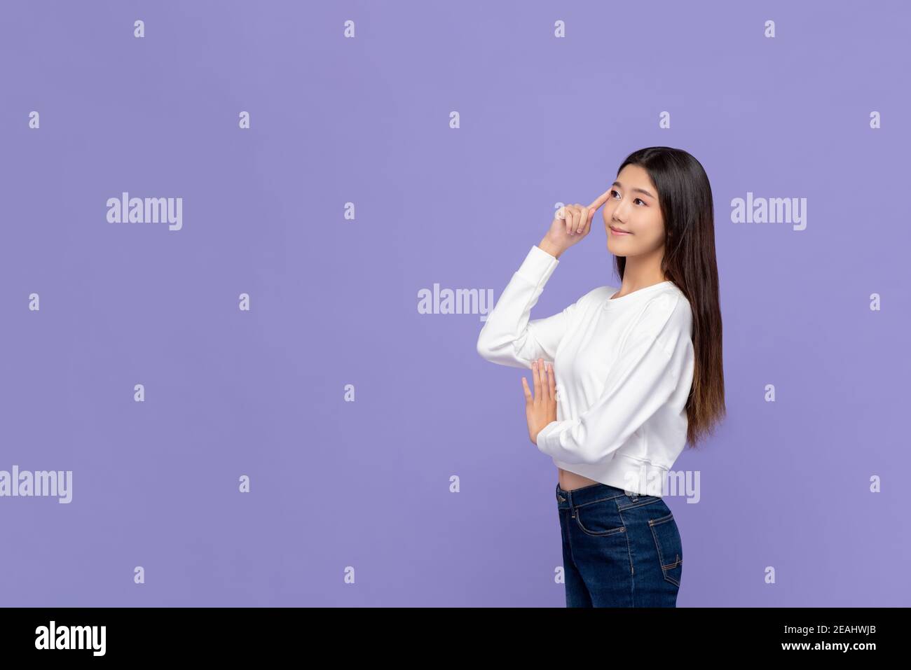 Attraente giovane donna asiatica sorridente che pensa con la testa che tocca la mano e guardando via lo spazio vuoto da parte su sfondo viola Foto Stock