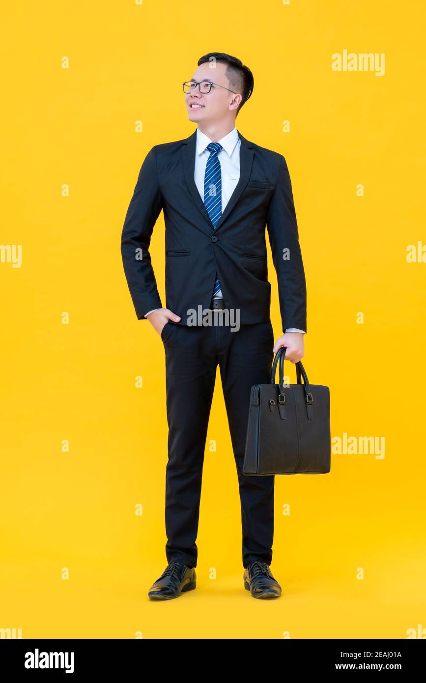 Felice uomo asiatico fiducioso in fomral tuta di affari in piedi e. testa di sollevamento isolata su sfondo giallo Foto Stock