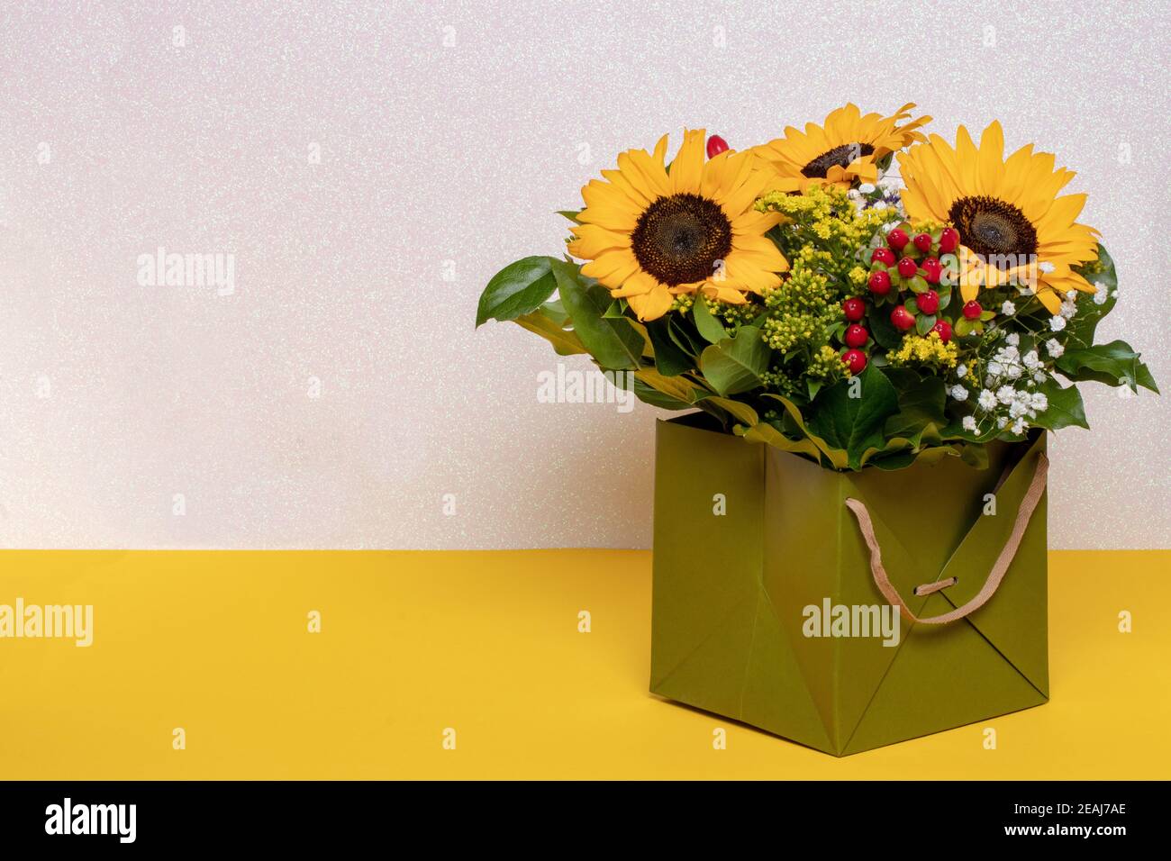 Closeup di un bel bouquet con girasoli gialli in una scatola regalo decorativa verde su un tavolo giallo su uno sfondo rosa chiaro scintillante. Macro. Spazio per il design. Concetto di scheda. Foto Stock
