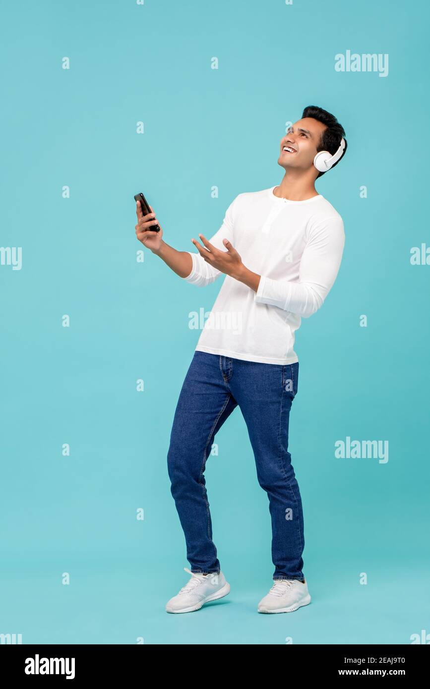 Sorridente giovane indiano in una maglietta bianca che ascolta la musica utilizzo di cuffie bluetooth wireless che tengono lo smartphone isolato in mano retro blu Foto Stock