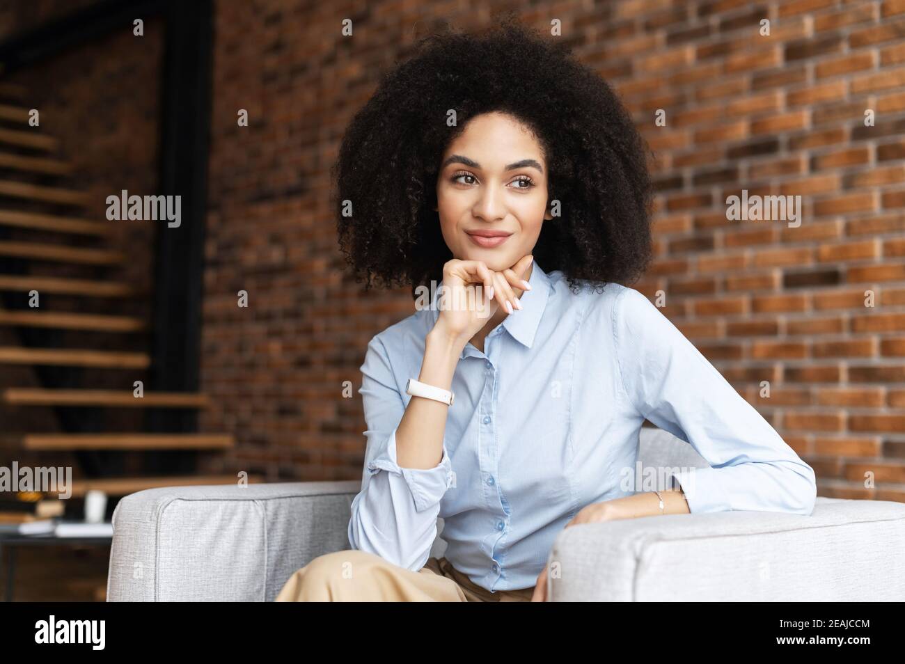 Un giovane imprenditore di razza mista sicuro di successo o una donna d'affari con capelli afro in abiti casual, seduto sulla sedia contro Foto Stock