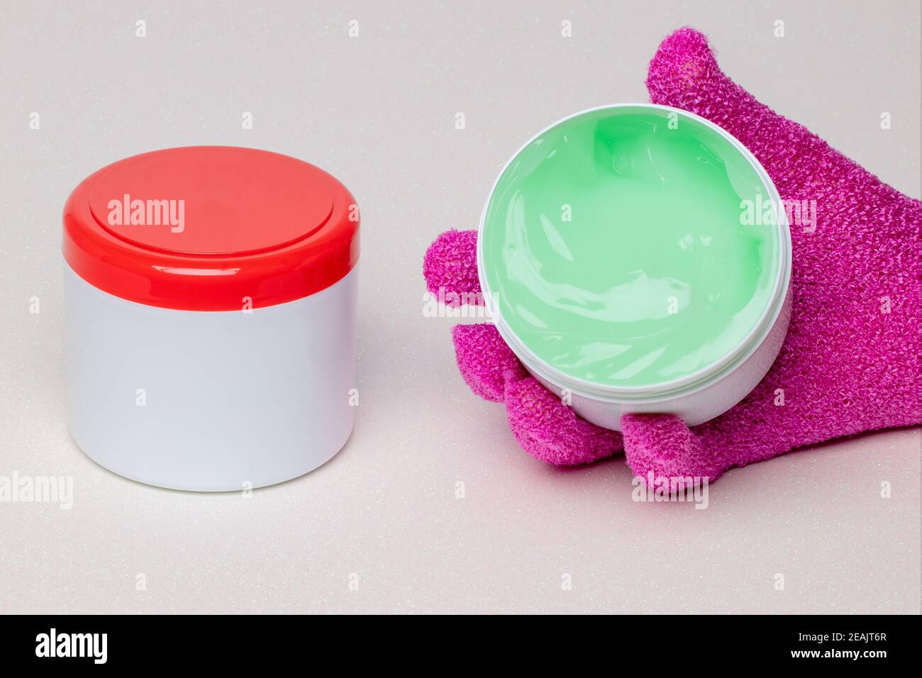 Chiusura di un contenitore chiuso e aperto con gel refrigerante (balsamo) per lesioni sportive su sfondo chiaro. Il contenitore aperto è tenuto da un guanto da massaggio rosa. Spazio di copia. Macro. Foto Stock