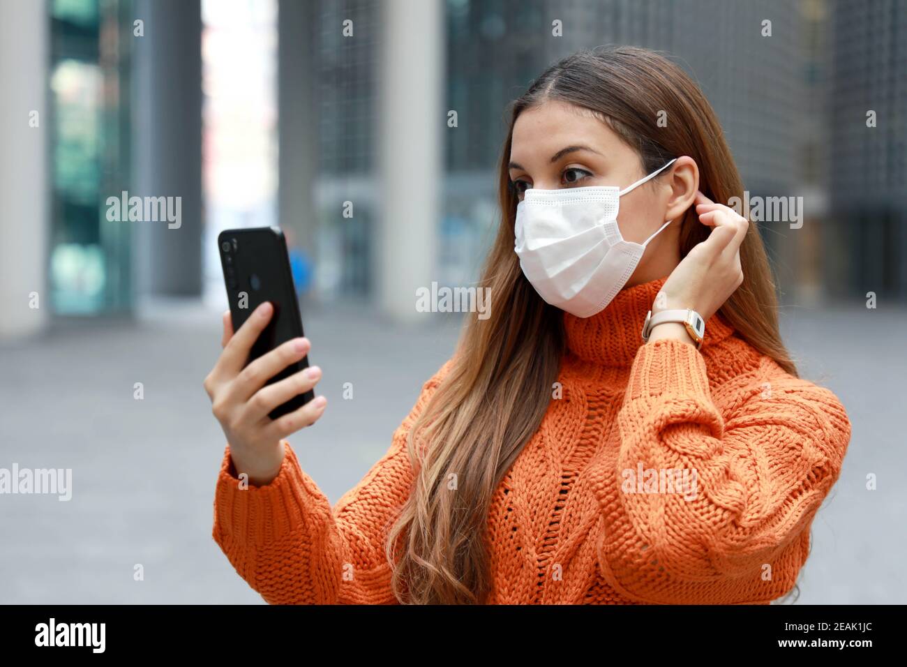 Videochiamate e Social Distancing. Bella giovane donna in città strada indossare maschera protettiva utilizzando il telefono cellulare per fare videochiamate o videoconferenza all'aperto. Foto Stock