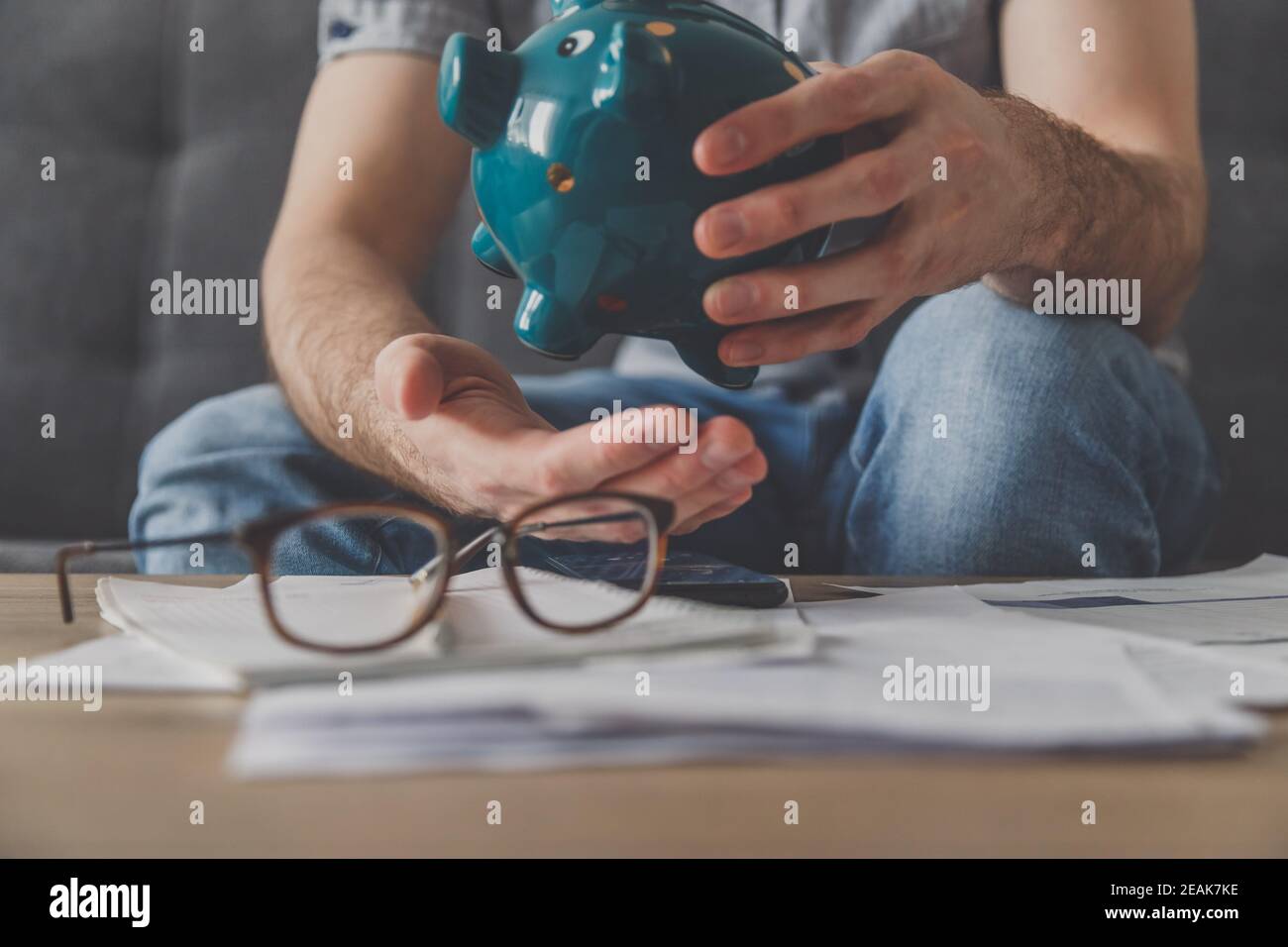 L'uomo seduto ad un tavolo pieno di fatture non pagate scuote fuori l'ultimo centesimo dalla banca piggy. Spendi i tuoi ultimi risparmi. Disoccupazione, povertà, concetto fallimentare. Foto Stock