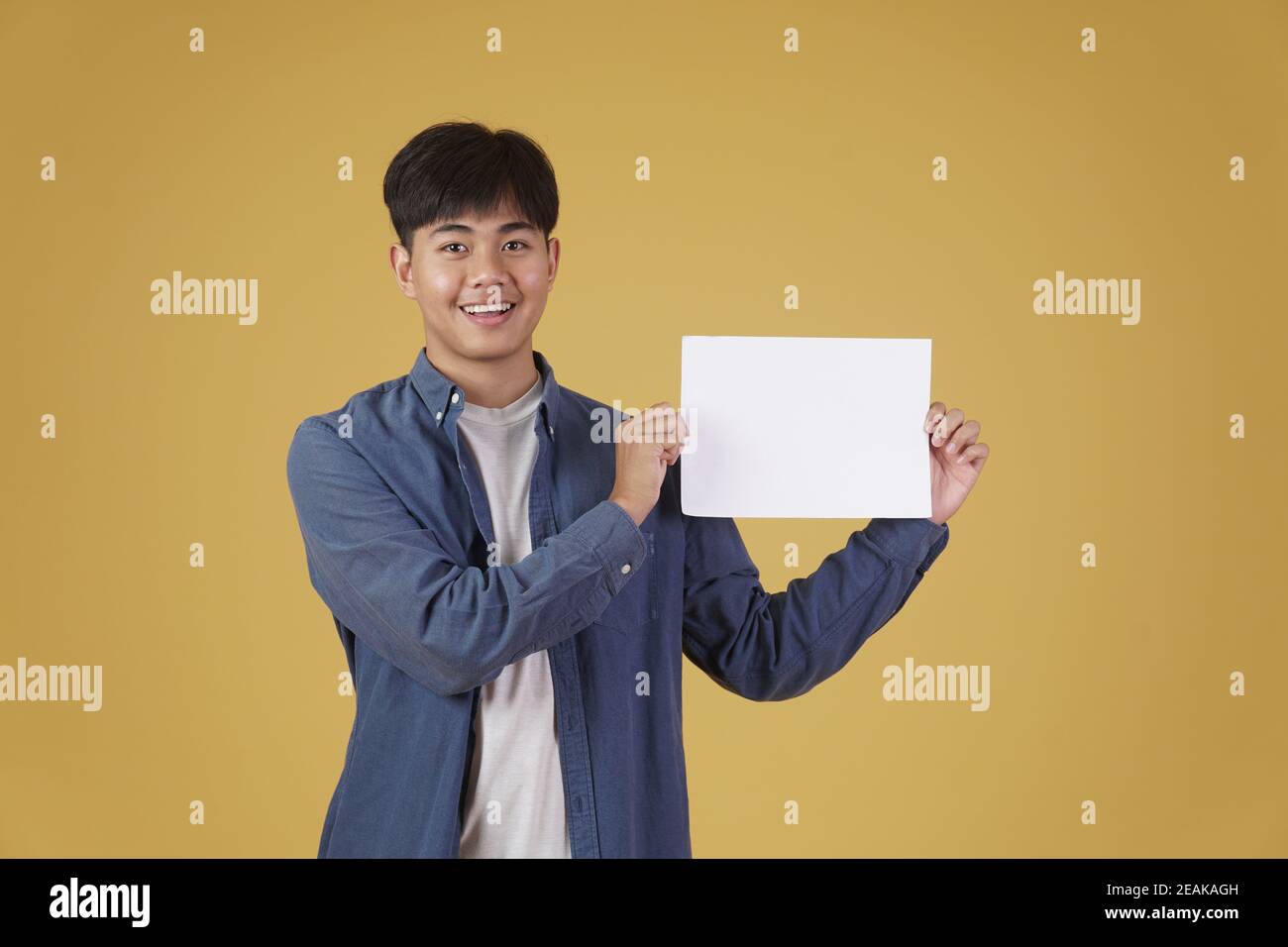 ritratto di sorridente felice allegro giovane asiatico vestito casualmente mostrando carta vuoto cartello isolato su sfondo giallo studio. Foto Stock