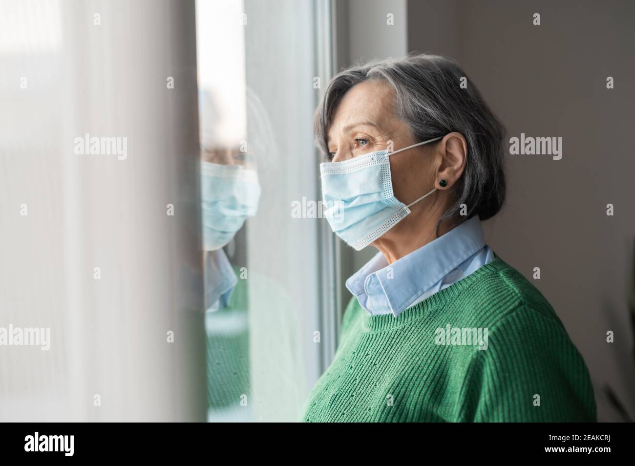 Una donna anziana con capelli grigi di media età che indossa un pullover verde e una protezione maschera facciale guardando la finestra e sentendosi triste depresso e solo Foto Stock