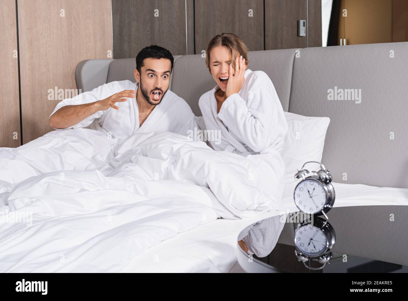 La coppia interracial stressata in accappatoi seduti sul letto vicino all'allarme orologio in primo piano sfocato in hotel Foto Stock