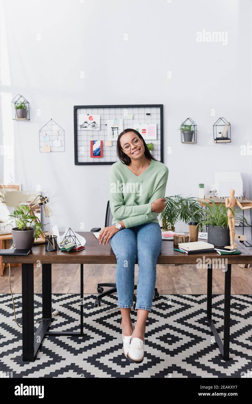 sorridente designer di interni afroamericano seduto sulla scrivania a casa studio e guardando la fotocamera Foto Stock