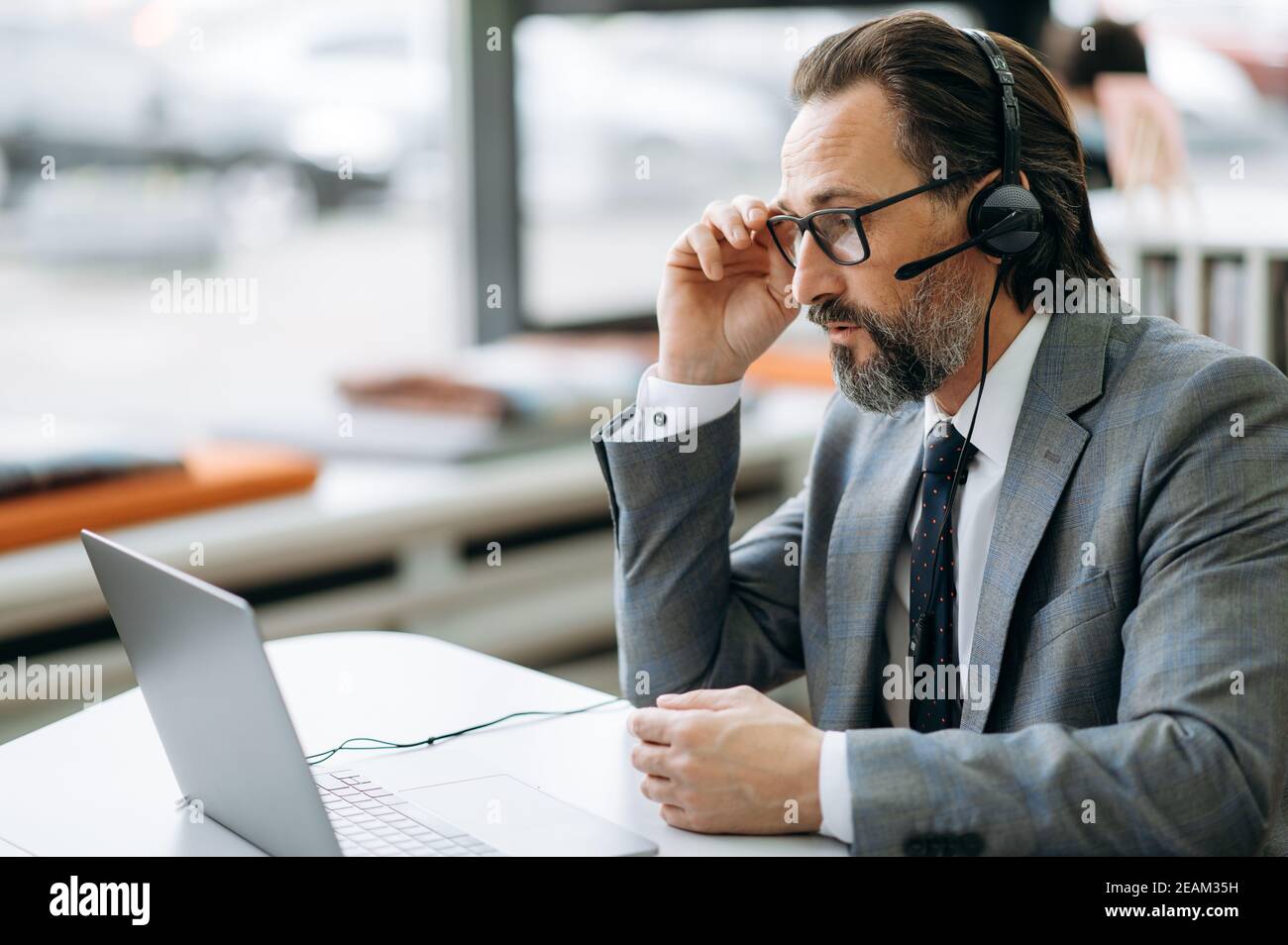 L'operatore maschile mirato del call center o del ceo è seduto alla scrivania e ha una conversazione seria. L'uomo d'affari senior indossa occhiali, tuta formale e cuffie comunicare online Foto Stock