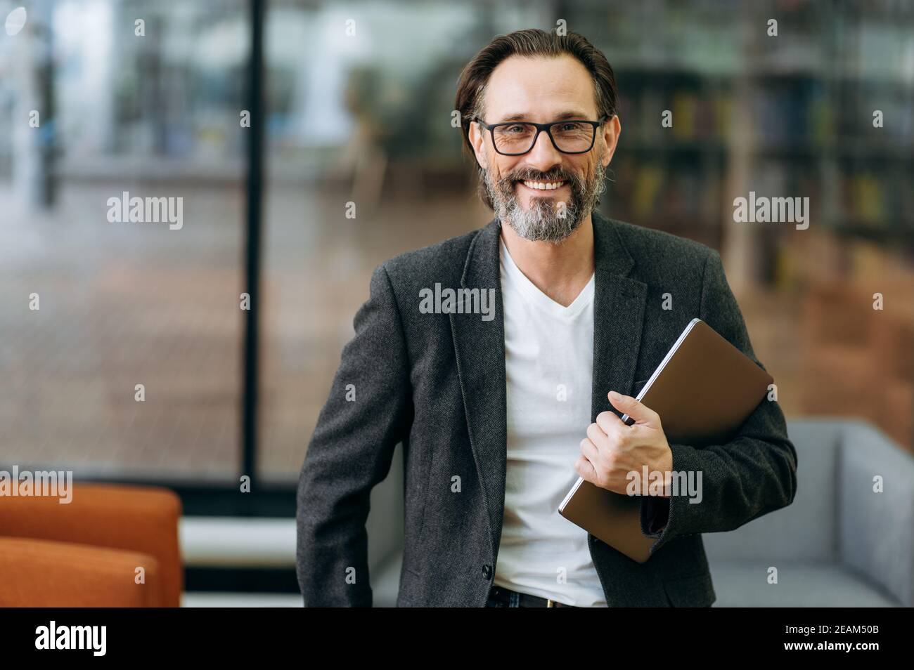 Ritratto di uomo d'affari felice sicuro in occhiali e abito formale elegante. Medio vecchio leader di successo maschile di affari guardando direttamente la fotocamera, sorridendo, tenendo il laptop in braccio Foto Stock