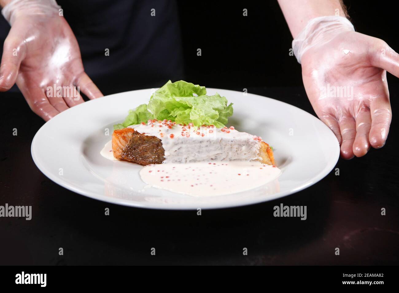 Le mani dello chef servono piatti da un pezzo di salmone in una cremosa salsa caviale su un piatto bianco con foglie di insalata. Foto macro. Foto Stock